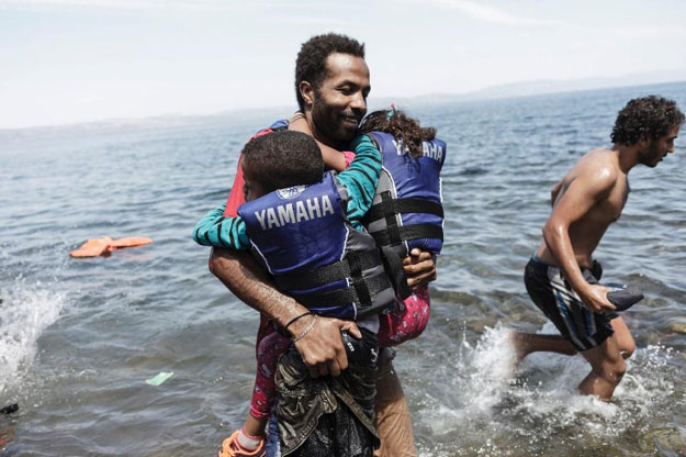 a migrant carries his two children as he gets off an inflatable boat after arriving on the greek island of lesbos after crossing the aegean sea from turkey on august 14 2015 photo afp