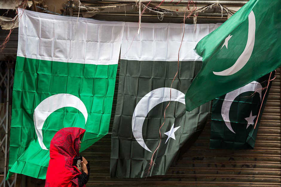 national flags in different hues of green are displayed against a wall as a woman walks past draped in a striking red dupatta photo zoral naik