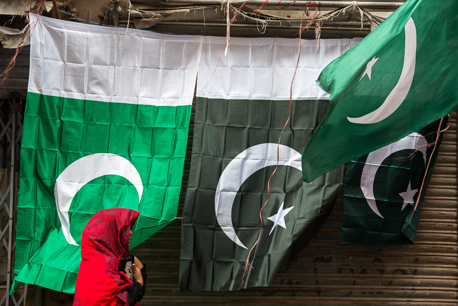 national flags in different hues of green are displayed against a all as a woman walks past draped in a striking red dupatta