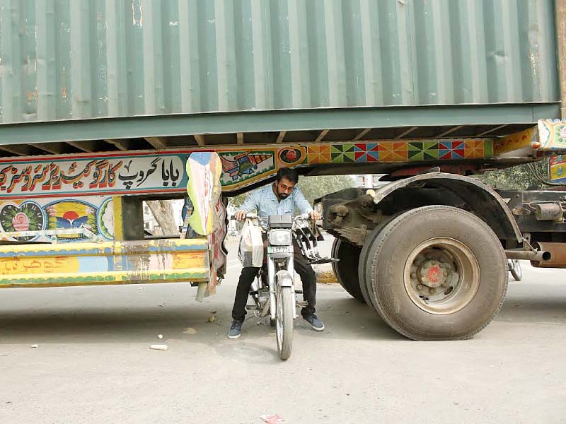a motorcyclist tries to squeeze through from under a truck used to block a road going towards a protest held by farmers on mall road photo abid nawaz express