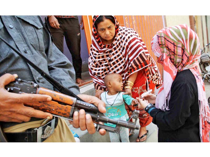 a policeman stands alert with his smg as a vaccinator marks the thumb of a child after administering drops of oral polio vaccine on the second day of anti polio drive in rawalpindi photo online