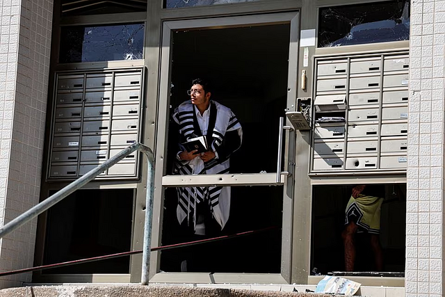 man wearing a Jewish prayer shawl looks out of the damaged entrance way to a building, as rockets are launched from the Gaza Strip, in Ashkelon, southern Israel October 7. REUTERS