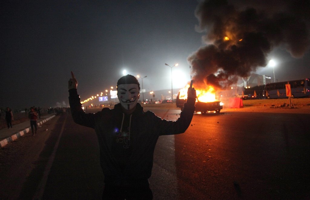 an egyptian man wearing a mask of the anonymous movement gestures near a burning car outside a sports stadium in a cairo 039 s northeast district on february 8 2015 during clashes between supporters of zamalek football club and security forces photo afp