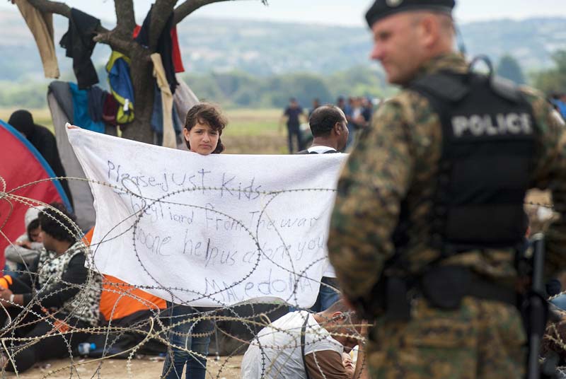 a police officer stand by near a young girl from syria holding up a towel reading quot please just kids tired from the war no one help us alone you macedonia quot as migrants wait behind barbed wire in a no man 039 s land between greece and macedonia to cross the border into the macedonian town of gevgelija on august 22 2015 photo afp