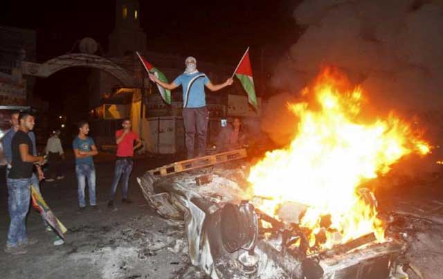 a palestinian holding palestinian flags stands on a burning car belonging to jewish settlers after it was set on fire by palestinians in the west bank city of nablus october 18 2015 photo reuters