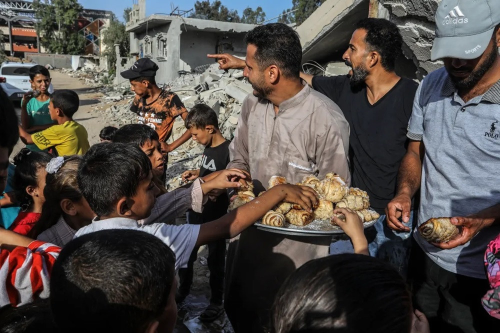 A Palestinian man distributes sweet buns on the Eidul Azha morning among the rubble of buildings destroyed by Israeli attacks in Khan Younis. PHOTO: ANADOLU AGENCY