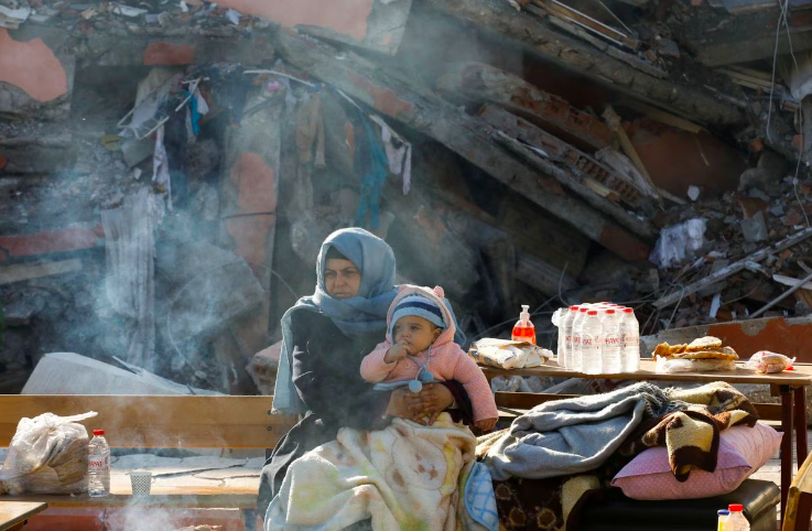 A woman holding a child sits by a collapsed building as search for survivors continues, in the aftermath of a deadly earthquake in Hatay, Turkey, February 10, 2023. Photo: REUTERS/Umit Bektas