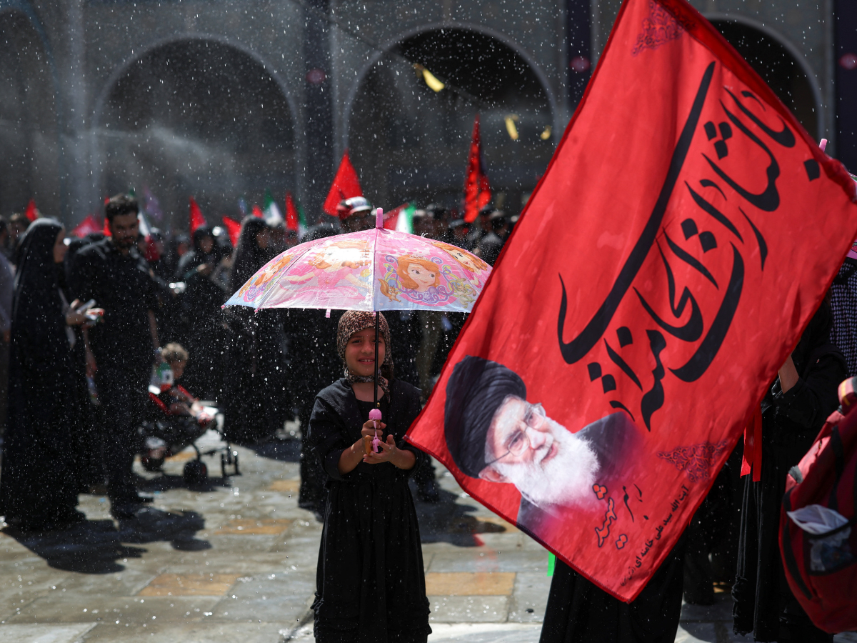 A girl stands under an umbrella as people cool themselves under sprayed water as they attend a public farewell ceremony to pay their respects to late Supreme Leader Ayatollah Ali Khamenei at the Imam Khomeini Grand Mosalla, in Tehran, Iran. Photo: Reuters