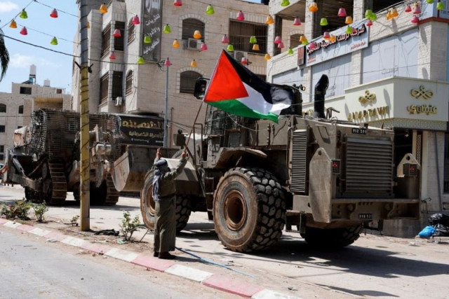 a palestinian man holds a national flag in front of israeli military vehicles during an israeli raid in the tulkarm refugee camp in the northern west bank on thursday august 22 2024 three palestinians were killed on thursday in an israeli bombing on a house in the tulkarm refugee camp according to the palestinian official news agency wafa photo xinhua a palestinian man holds a national flag in front of israeli military vehicles during an israeli raid in the tulkarm refugee camp in the northern west bank on thursday august 22 2024 three palestinians were killed on thursday in an israeli bombing on a house in the tulkarm refugee camp according to the palestinian official news agency wafa photo xinhua