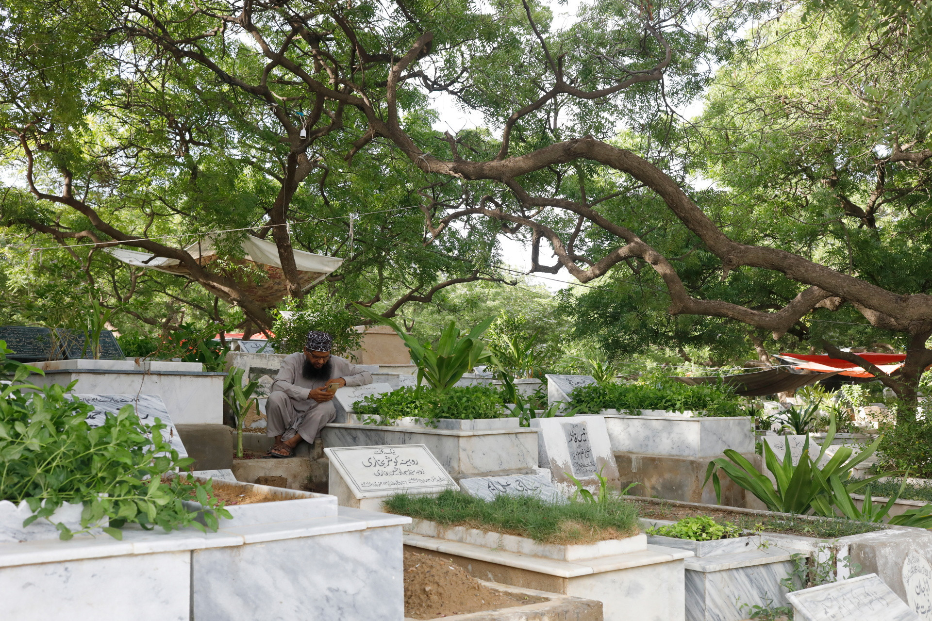 A man reads a prayer on a phone as he sits at Sakhi Hassan Graveyard, which is filled with plants seeded by relatives of the dead, in Karachi, July 12, 2022. REUTERS