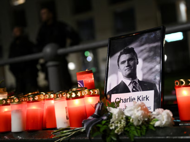Roses and candles are placed next to a picture of Charlie Kirk during a vigil under the line 